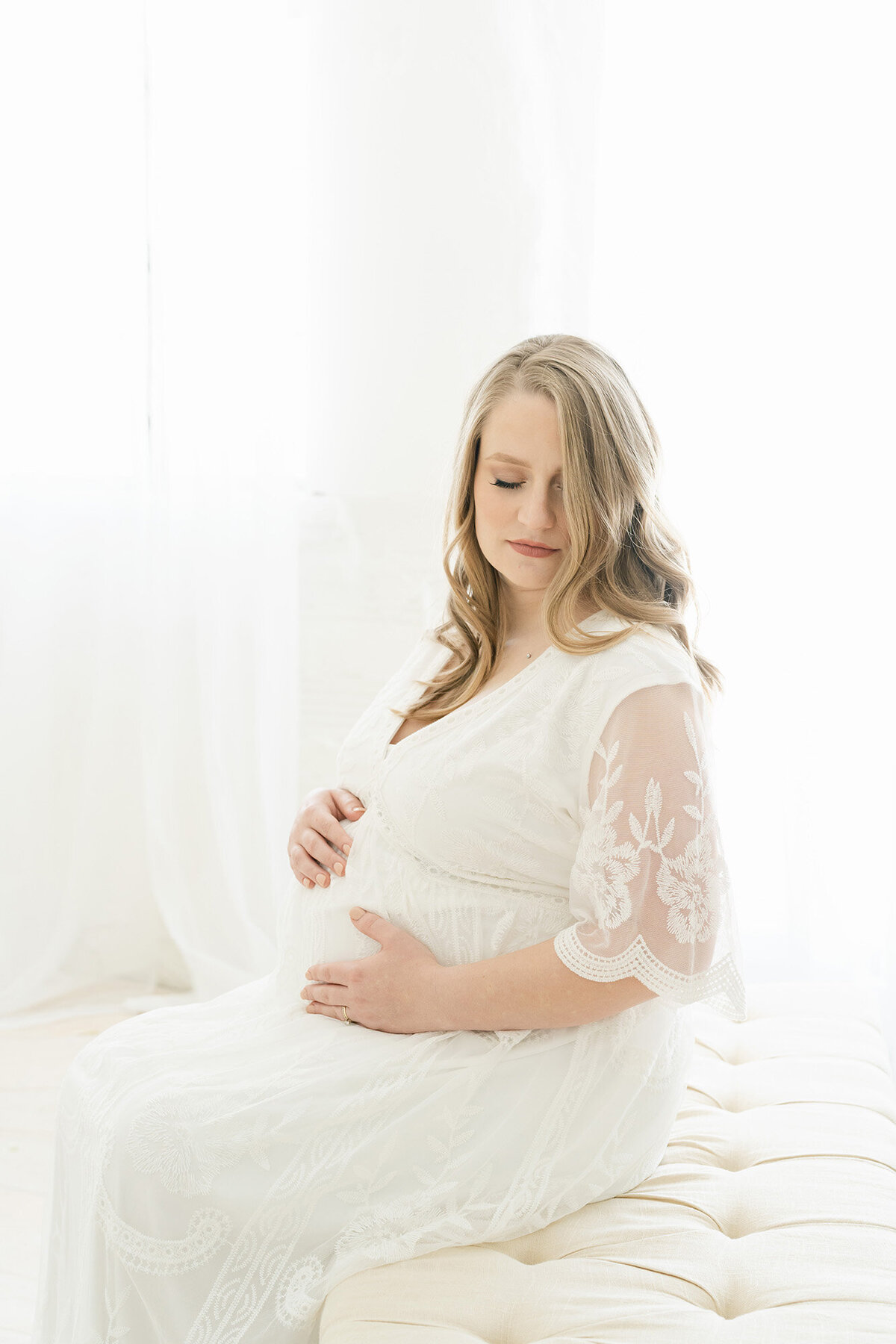 Pregnant mother sits on tufted bench and wears white dress at Louisville Ky photography studio