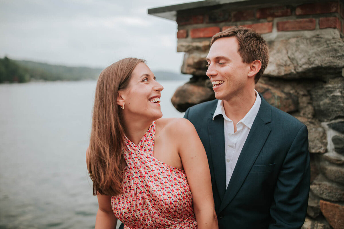 A smiling couple shares a joyful moment by a lakeside, with a rustic stone wall in the background.In Sparta NJ.
