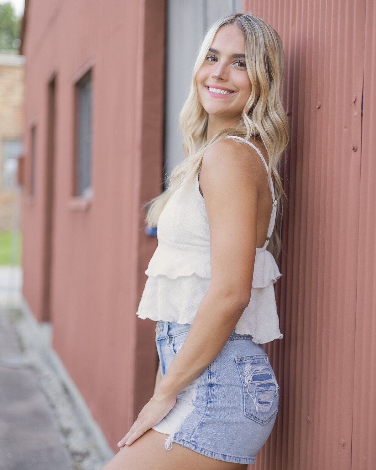 high school senior female with long blonde hair, standing against a dark red building, turned to the side smiling at the camera, wearing blue jean shorts, and white spaghetti strapped shirt