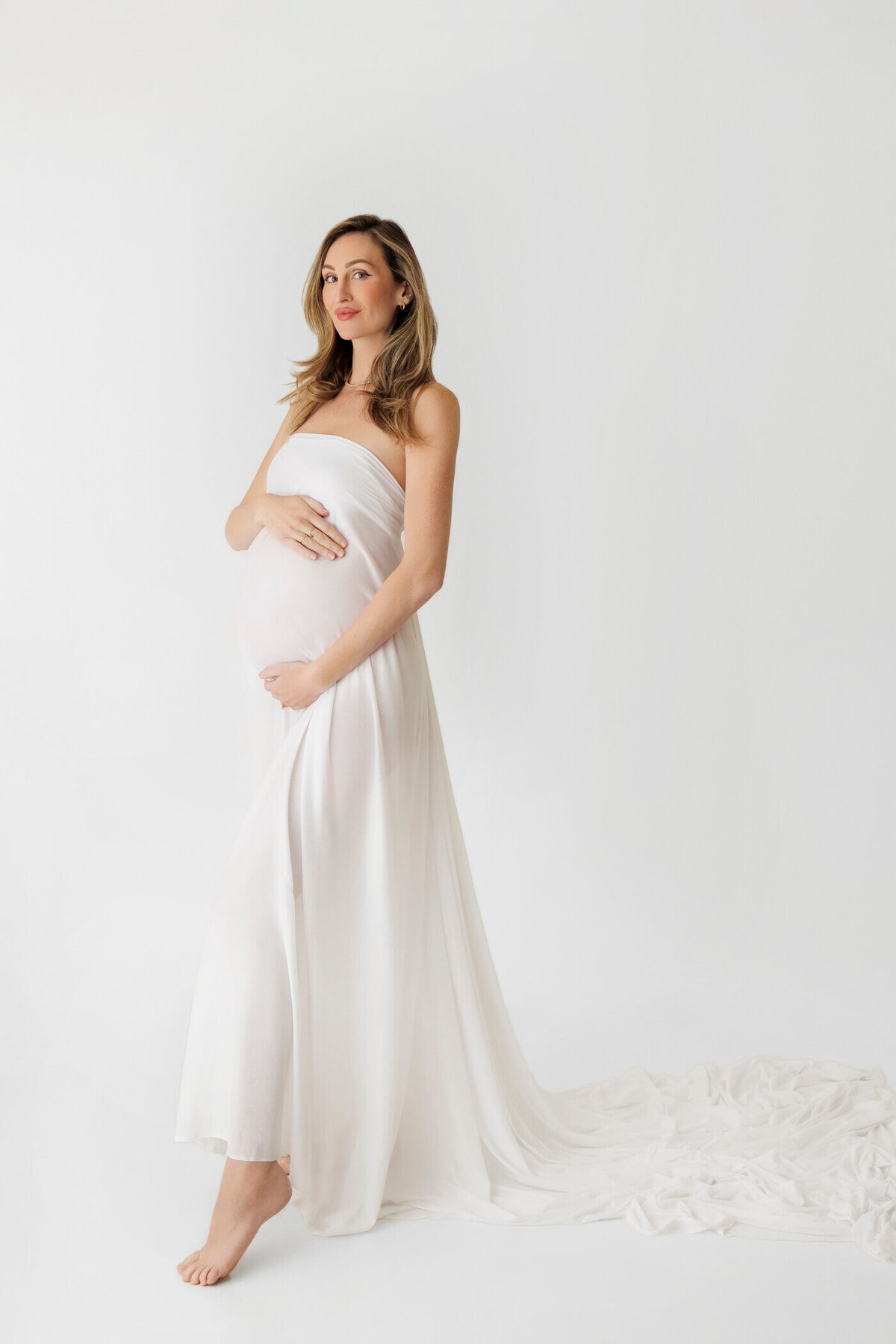 A pregnant woman stands gracefully barefoot against a pure white background, wrapped in a flowing, strapless white gown that drapes elegantly to the floor and pools behind her. She cradles her baby bump with both hands, one resting gently above and the other below. Her long hair cascades over her shoulders as she gazes softly into the distance, exuding a sense of calm and anticipation. The minimalist setting highlights the serene and ethereal beauty of pregnancy.