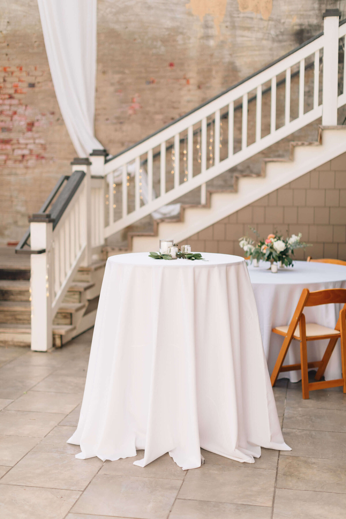 A round table with a white tablecloth holds a floral arrangement in a courtyard.