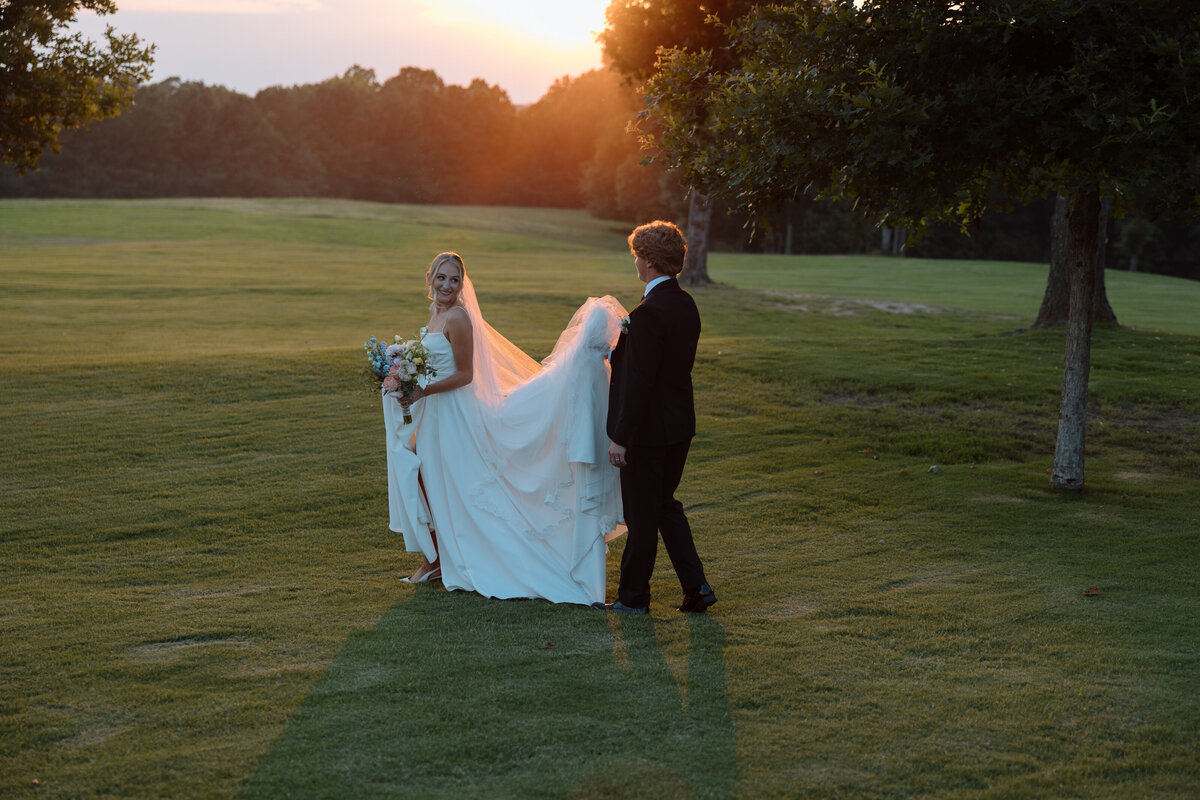 Bride and Groom walk through a field at sunset while groom holds the bride's dress at Thornblade Country Club in Greenville,SC