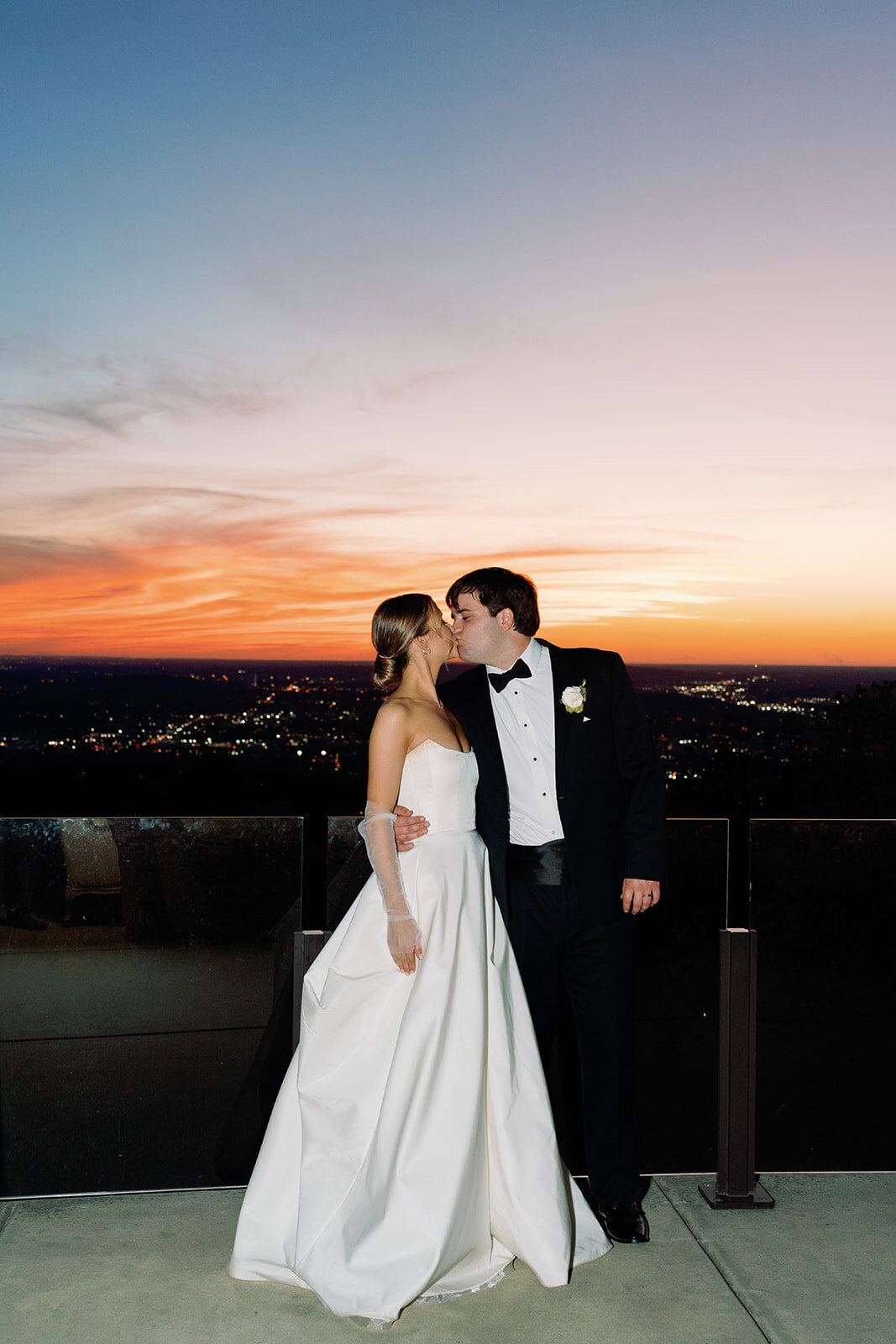 Bride and groom share a kiss on the terrace at Burritt on the Mountain with a vibrant sunset and city lights in the background.