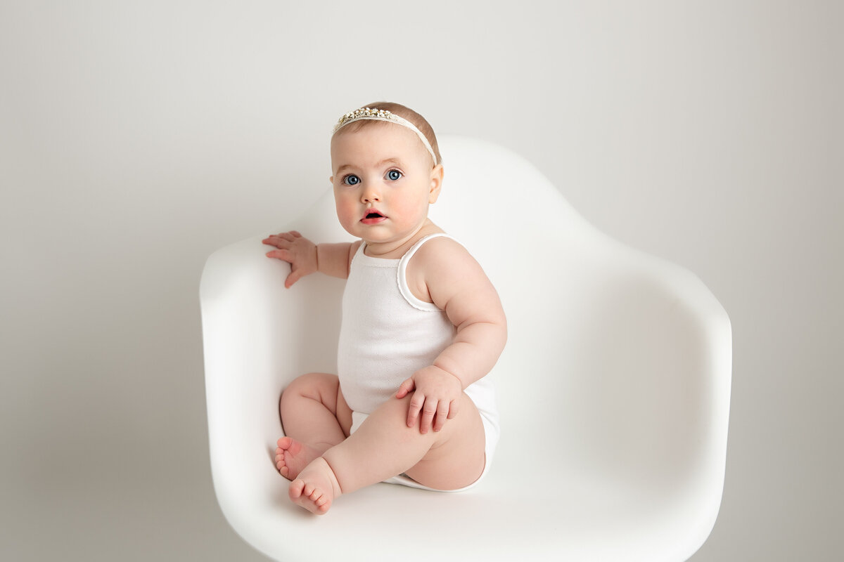 Six month old baby girl dressed in a white onesie and sitting on a white chair.