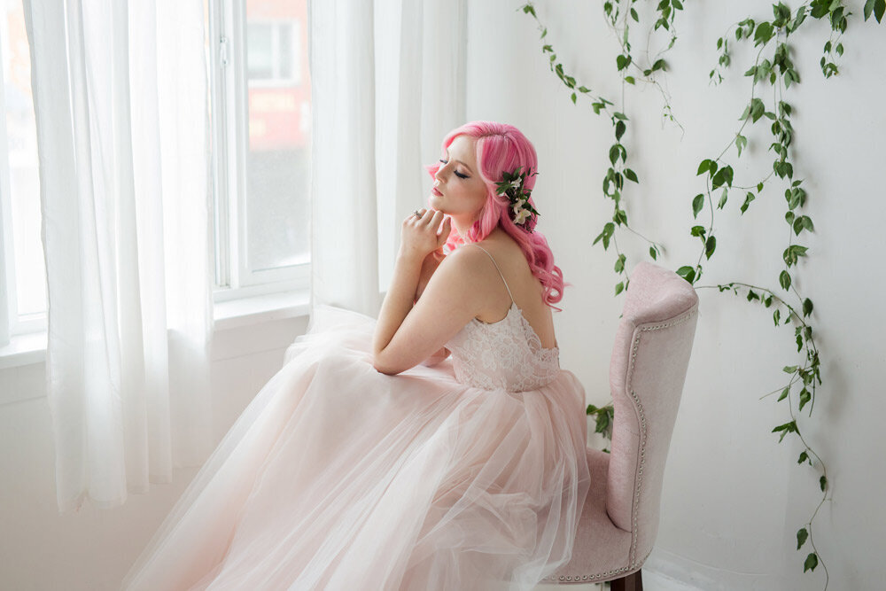 Bride with long, newly styled pink hair poses in a a chair while looking out window