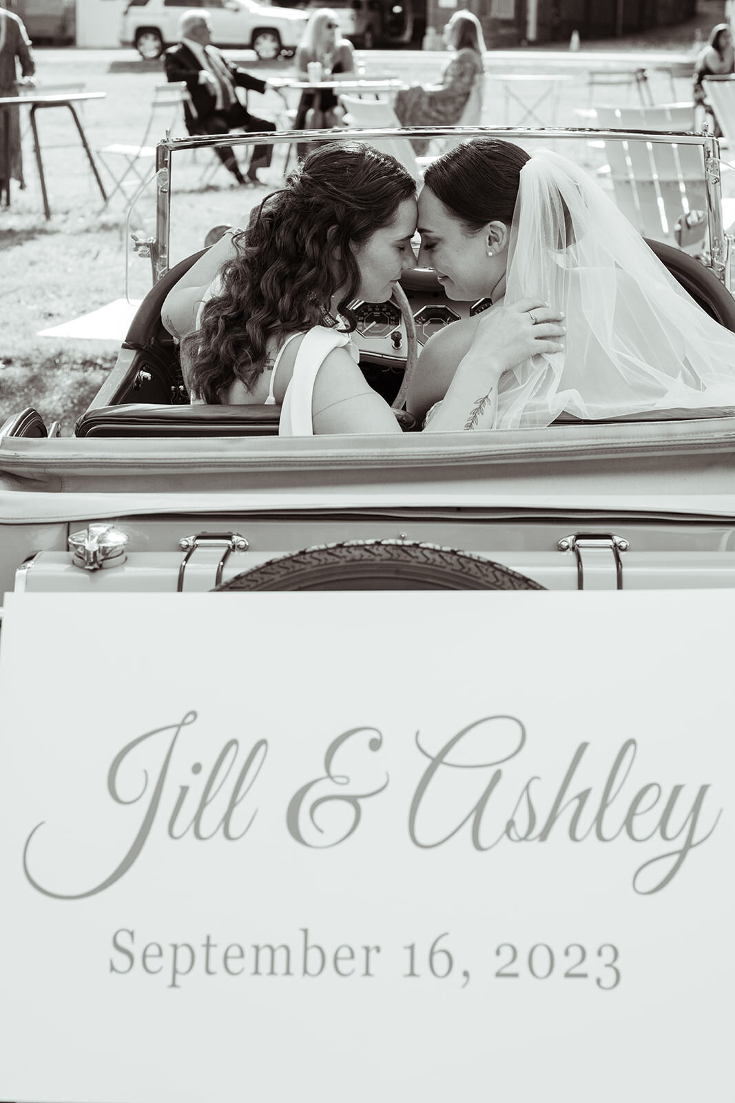 Jill & Ashley sitting in vintage car with a wedding sign with their names on it on the back