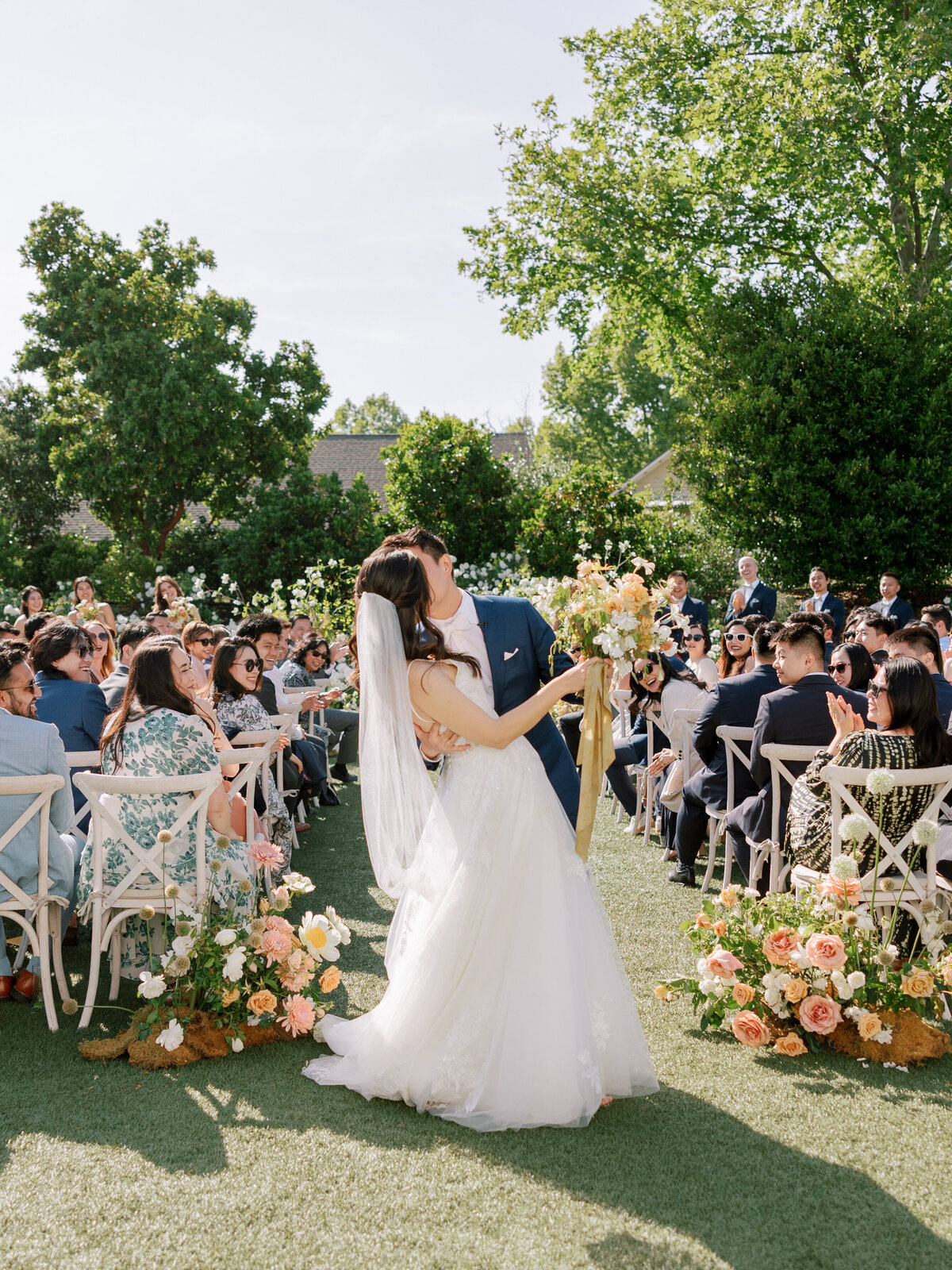 A couple sharing a celebratory kiss during their wedding ceremony recessional at Solage Napa Valley. Surrounded by smiling guests and the stunning wine country backdrop, this romantic moment captures the joy and love of their special day.