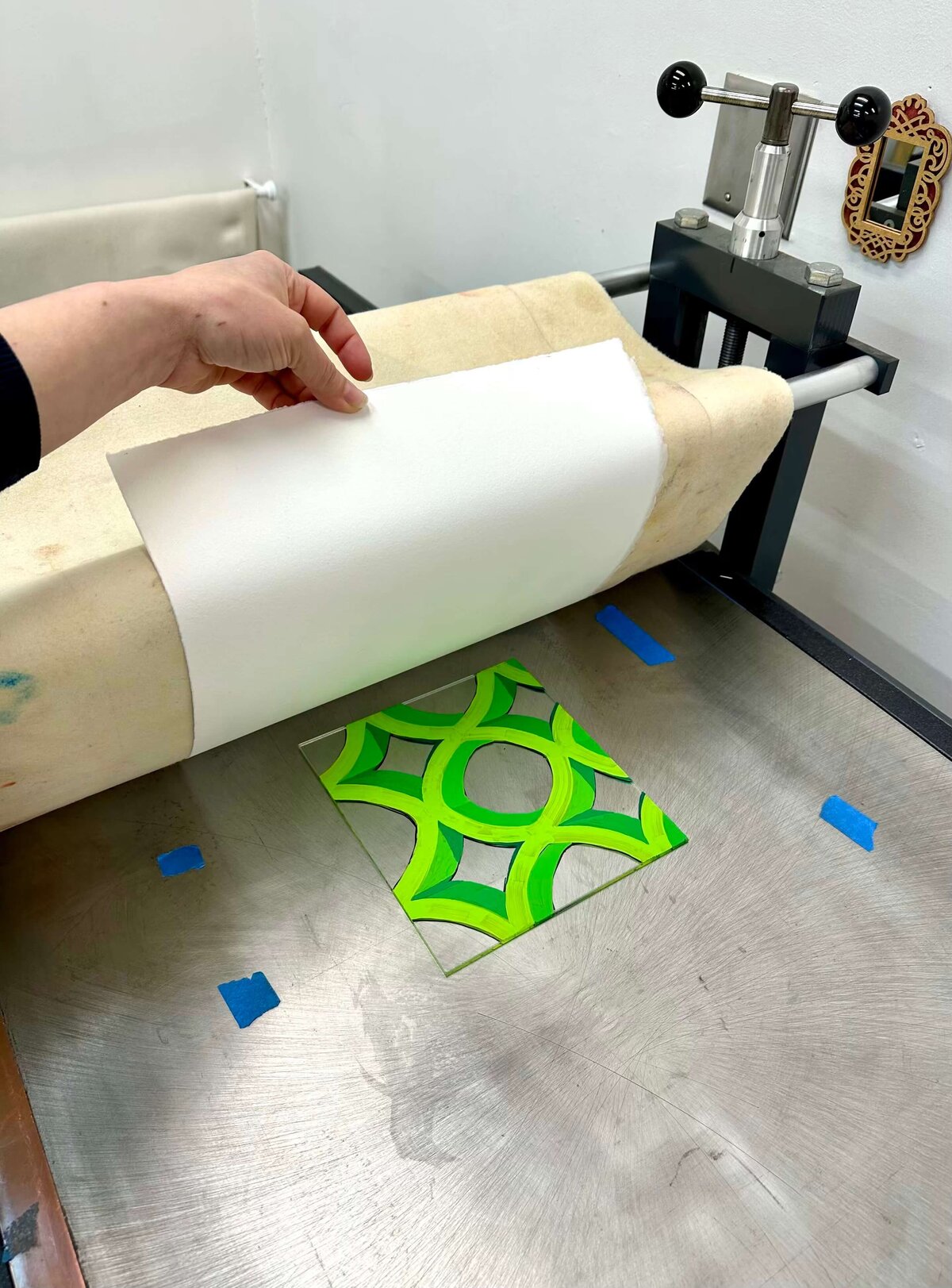 A green geometric design painted on plexiglass is prepped under an etching press with paper layered above, ready for printing.