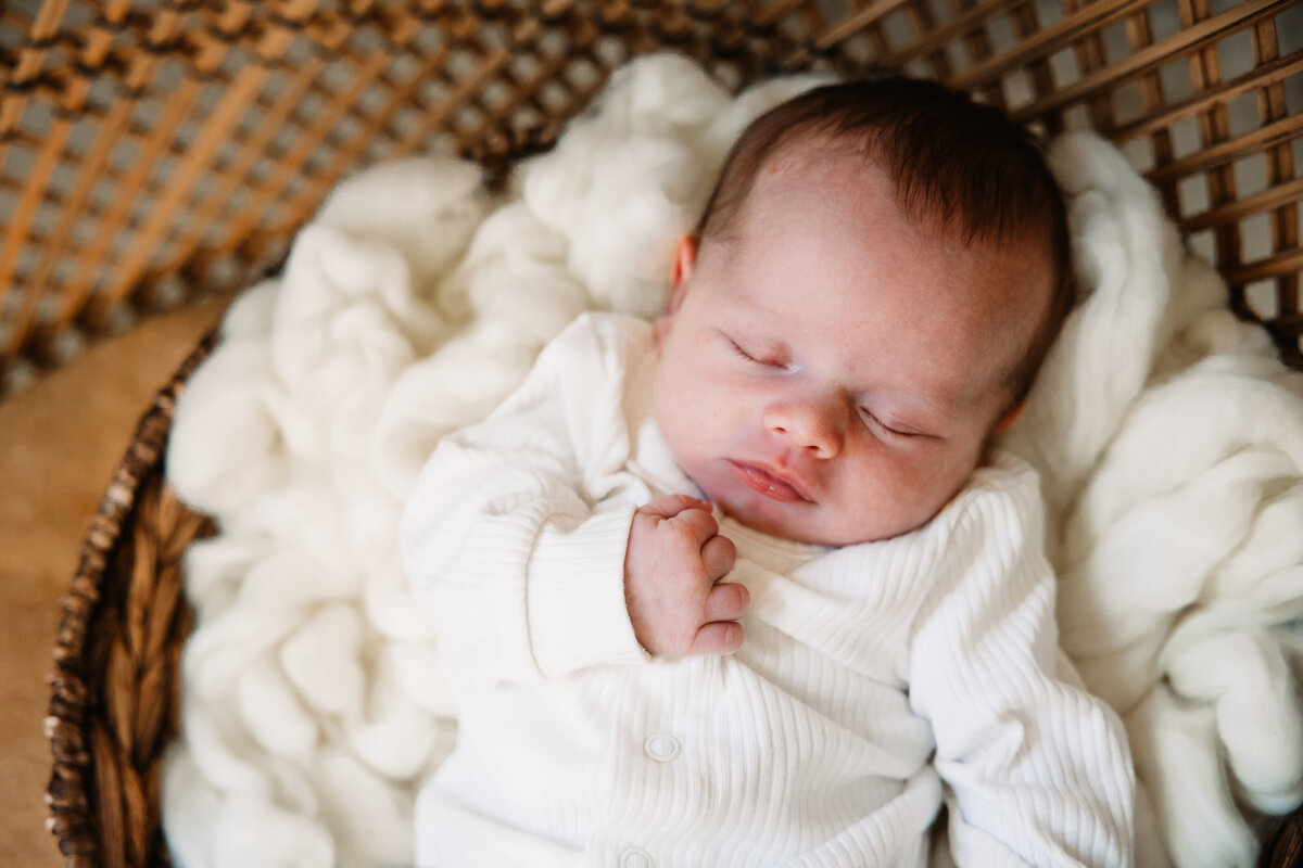 A newborn baby sleeps in a wicker basket wearing a white onesie