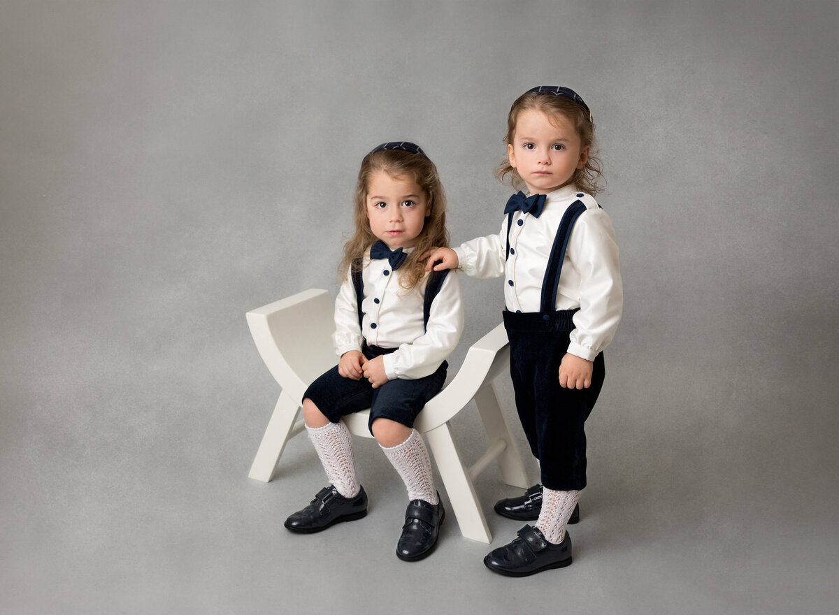 Toddler boys are posing for a milestone photoshoot. One boy is sitting in a chair, the other boy is standing with his hand on his brother's shoulder.upshernesh twins photos Nyc
