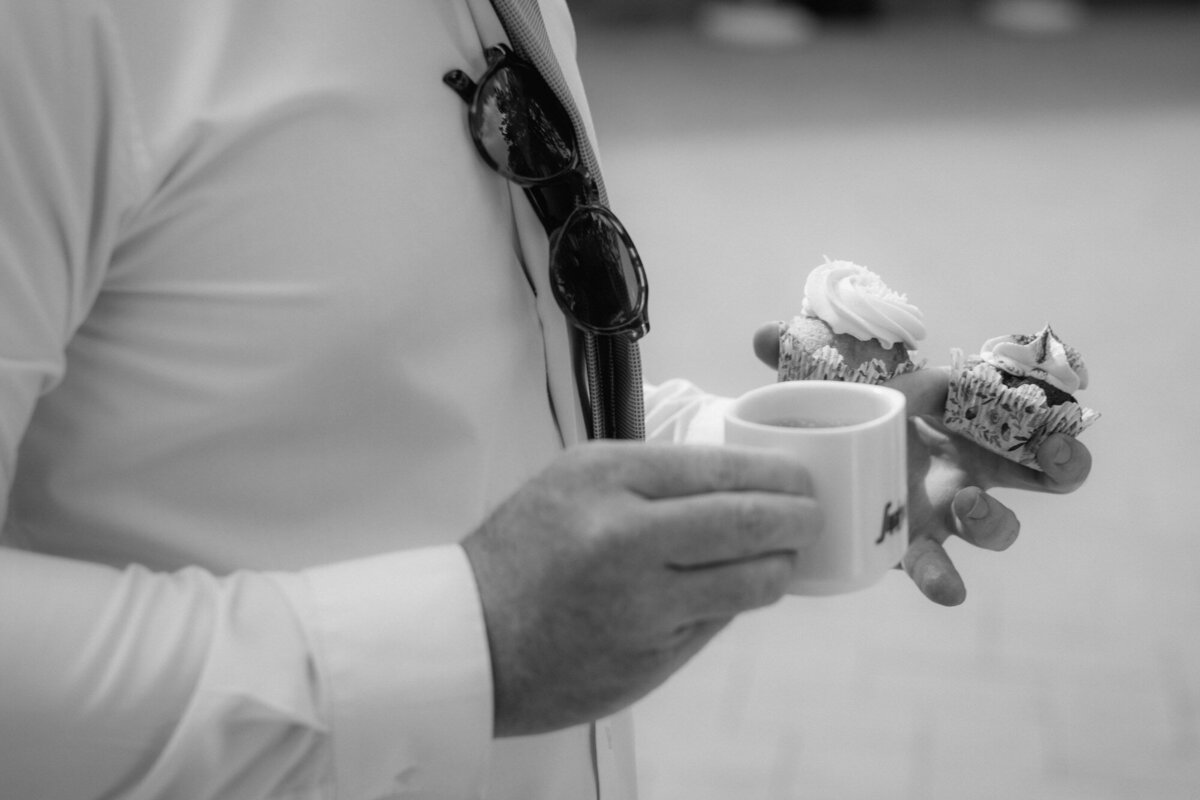 Ein männlicher Hochzeitsgast hält zwei Mini-Cupcakes und eine Kaffeetasse in der Hand.