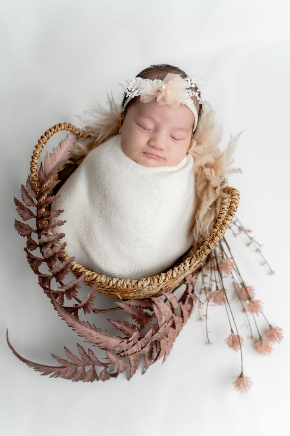 Newborn baby swaddled in a soft white wrap, wearing a delicate peach floral headband, sleeping in a wicker basket decorated with dried ferns.