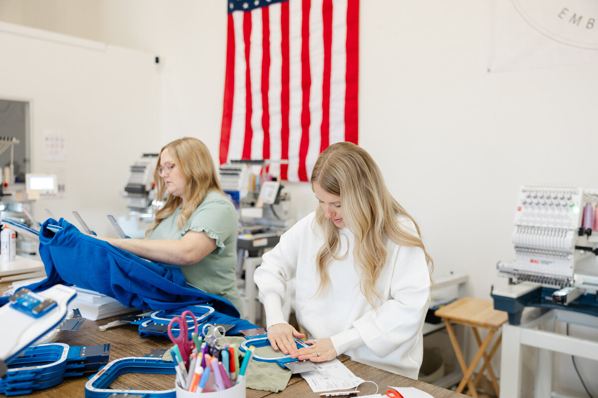 Two blonde women collaborating in embroidery studio with sewing supplies in background. Photograph by Yucaipa branding photographer Kaitlyn Dawn Photography.