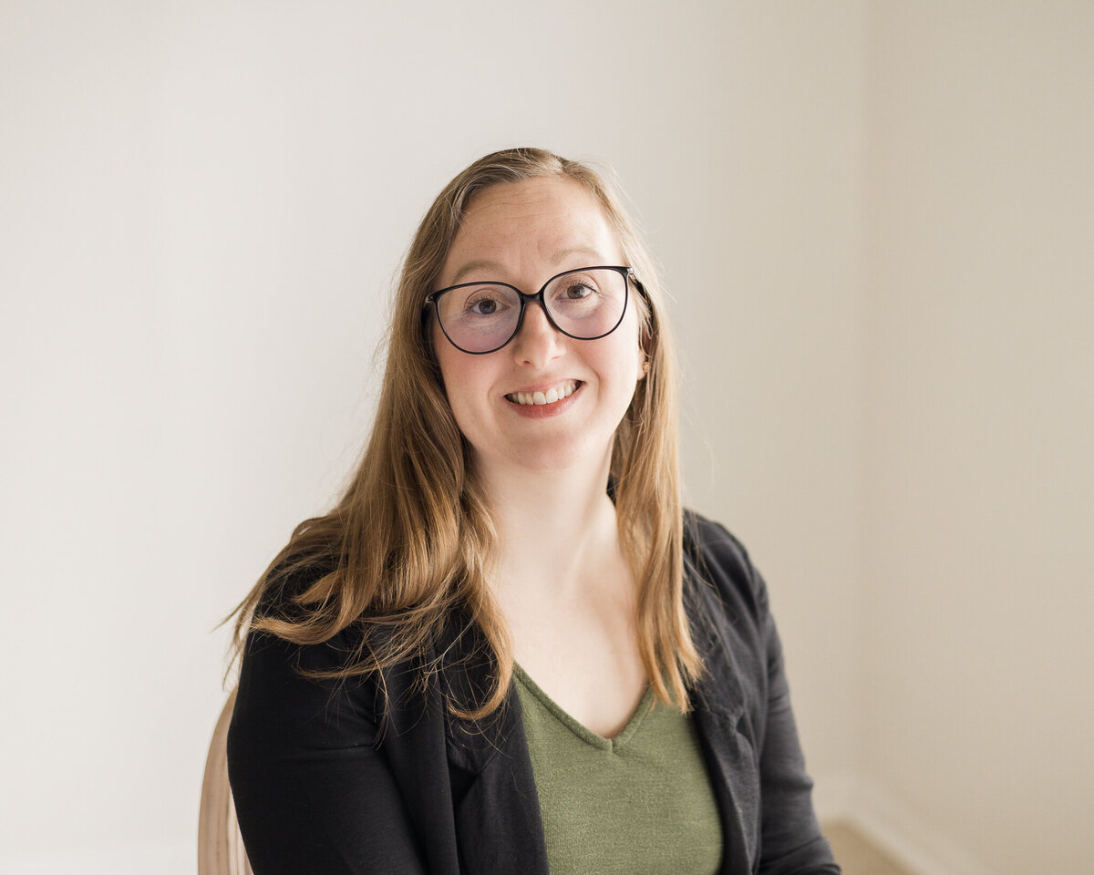 Strawberry blonde woman sitting in a chair, wearing glasses, a black dress jacket over a hunter green shirt and pants, wearing glasses, smiling at the camera