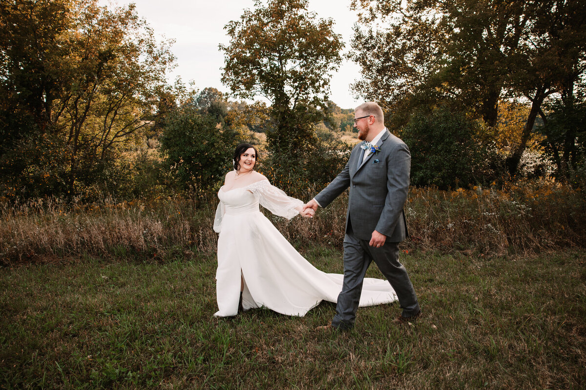 A bride leads her groom by the hand down a trail at sunset while smiling back at him