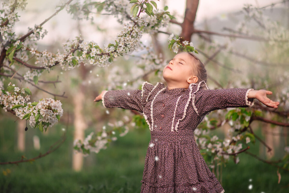 A little girl wearing a purple dress stands with her arms spread out wide and her eyes closed and head facing the sky. Portrait taken in Kamloops at a cherry orchard.