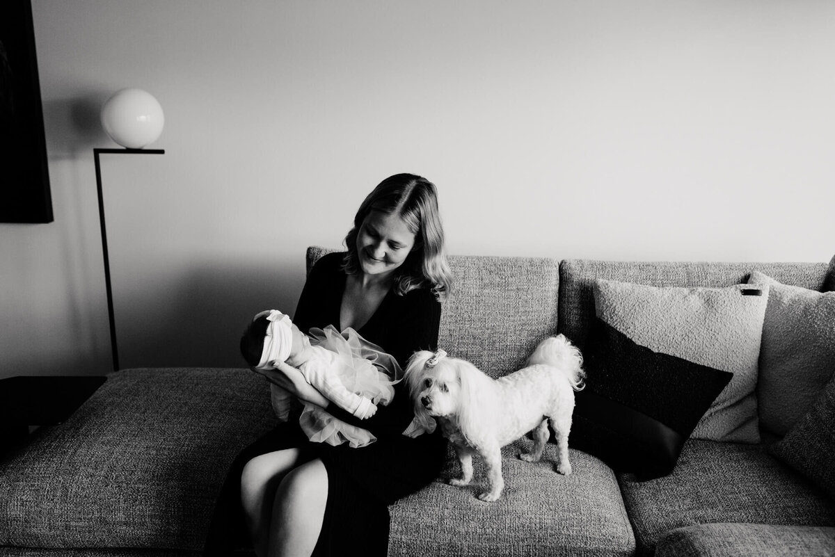 A black and white image of a mother sitting on her couch in a black dress holding her baby and sitting next to her little white dog