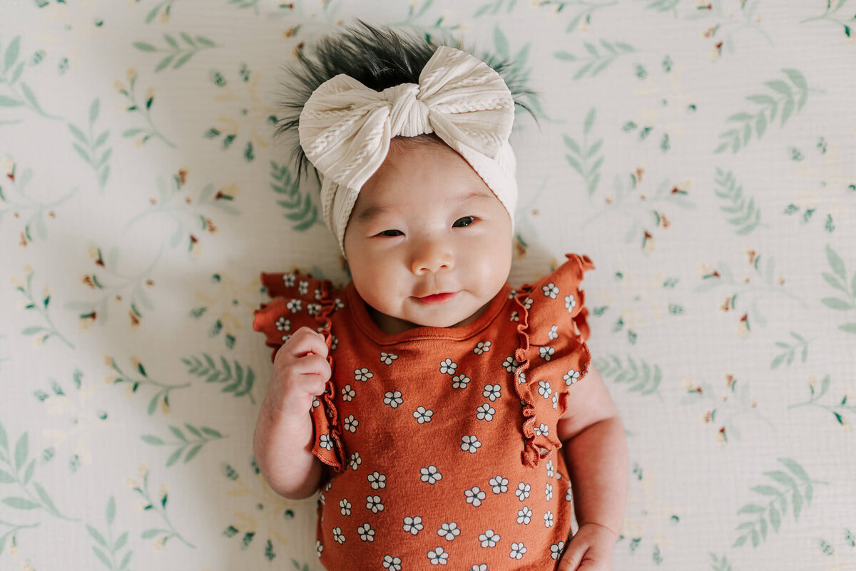 A newborn girl wearing a rust floral onesie and big neutral bow, taken by Denise Van Photography