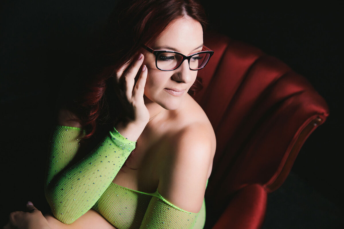 Darkly lit boudoir portrait of red haired woman in glasses and sheer green bodysuit