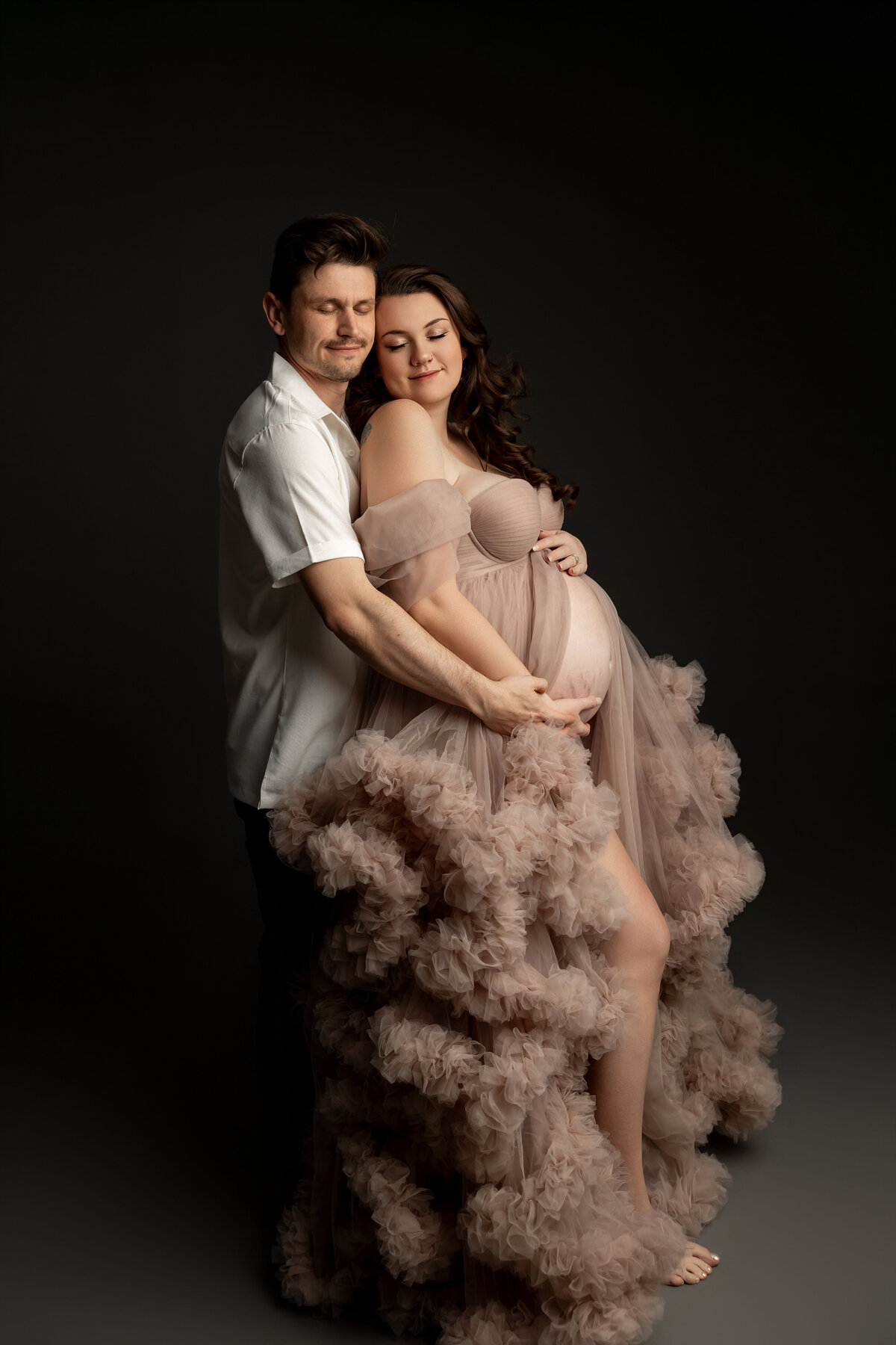 A beautiful and intimate maternity portrait featuring a glowing mother-to-be in a soft, ruffled taupe gown, lovingly embraced by her partner. Captured against a rich dark backdrop, this timeless image by Inland Empire maternity photographer Kaitlyn Dawn Photography perfectly showcases the love and anticipation of welcoming new life.