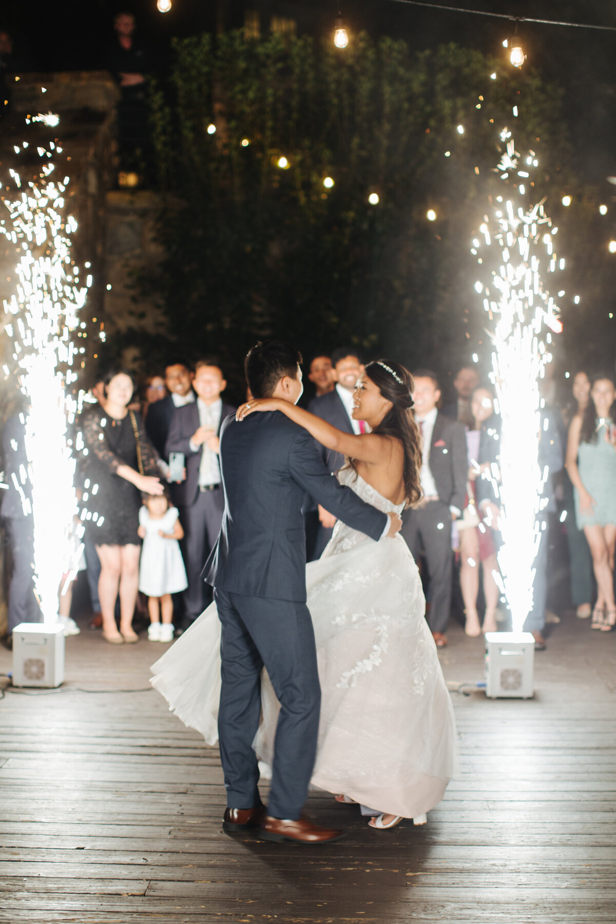 Couple dancing under string lights with cold spark fountains creating a dramatic entrance during their wedding reception.