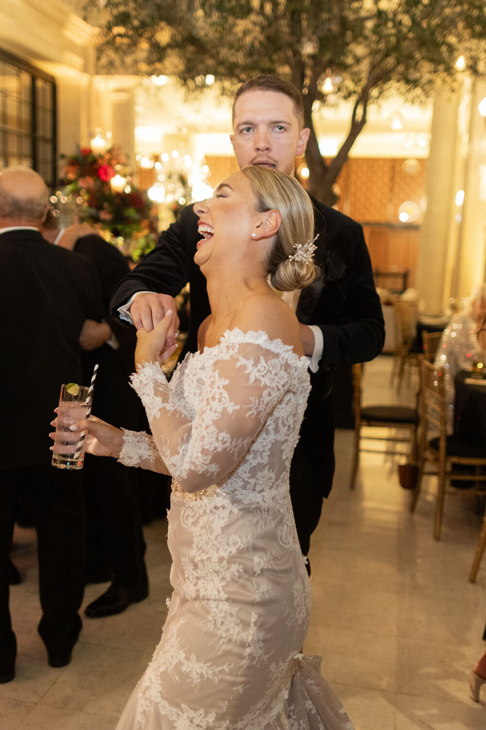 A woman in a white lace dress is laughing while holding a glass