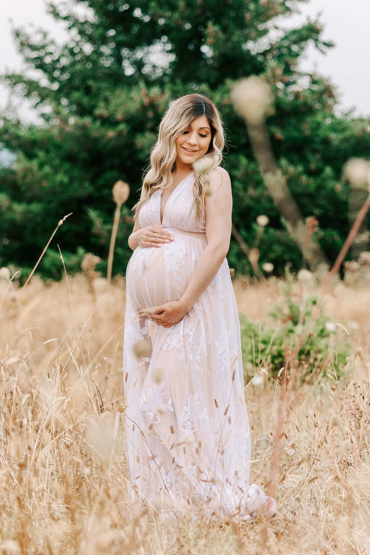Portrait of a woman in a field with tree behind her. She is looking over her shoulder and smiling while holding her pregnant belly..