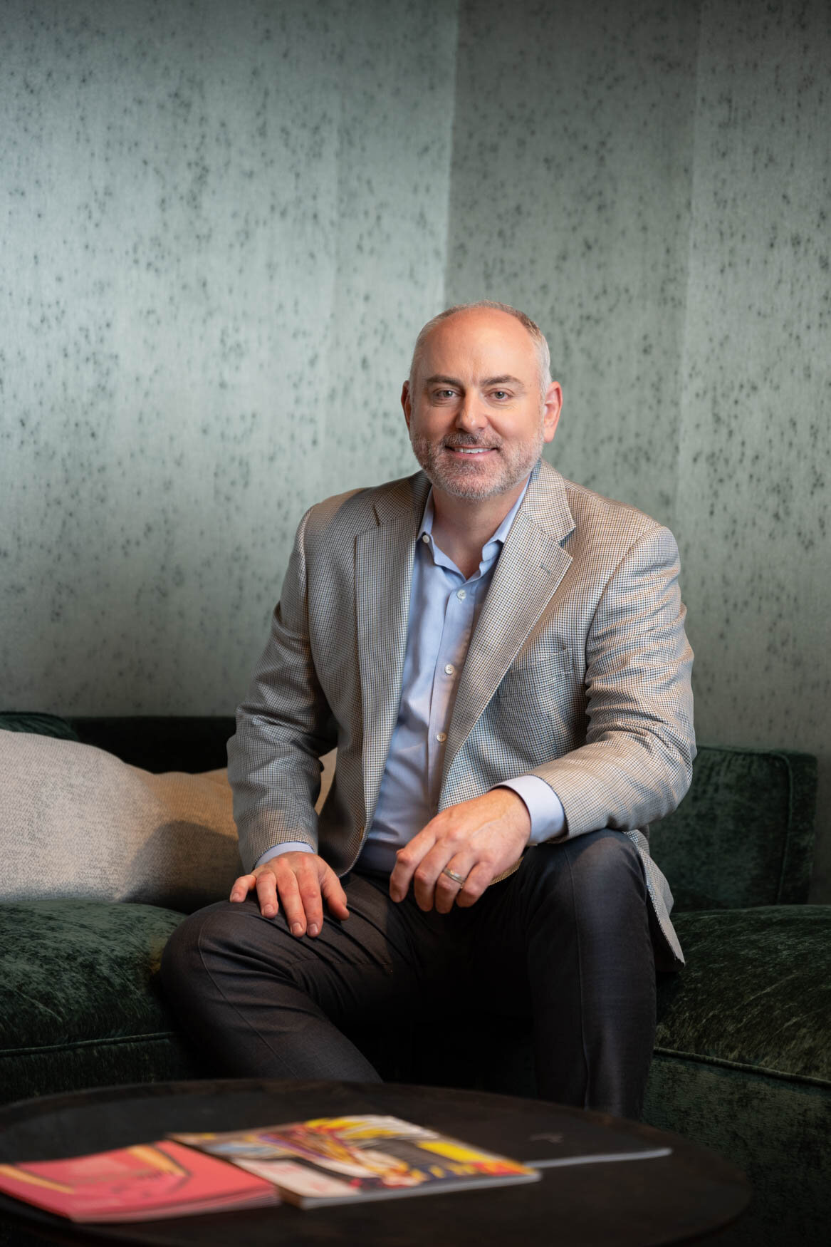 A man sitting on a green couch smiing to the camera with his arm on one of his legs