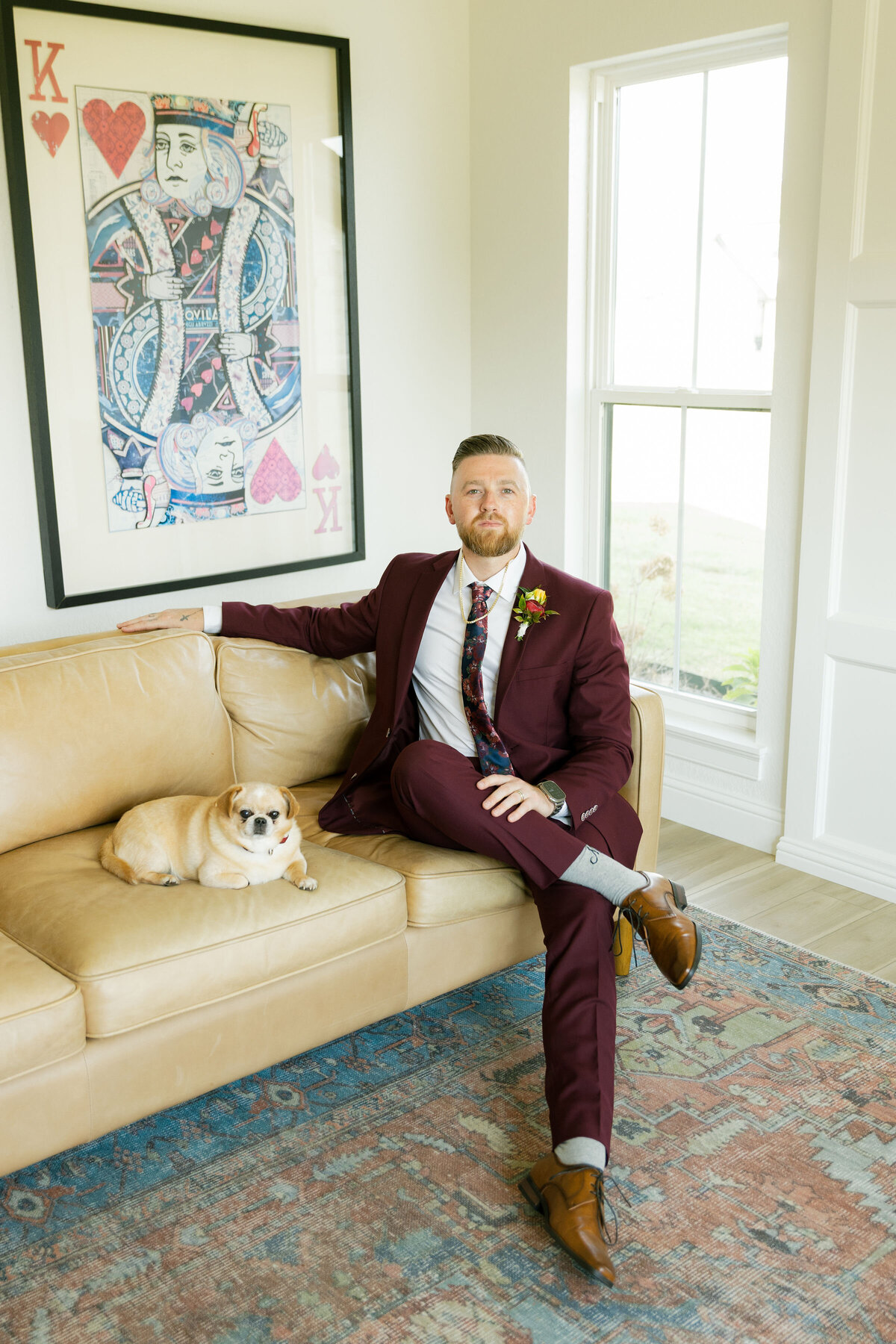 A groom dressed in a burgundy wedding suit sits confidently on a tan leather couch beside his small dog in a bright, modern room with large windows. Behind him hangs a large King of Hearts artwork, adding personality and style to the groom’s indoor wedding portrait. Warm tones, relaxed posing, and natural light create a polished editorial feel.
