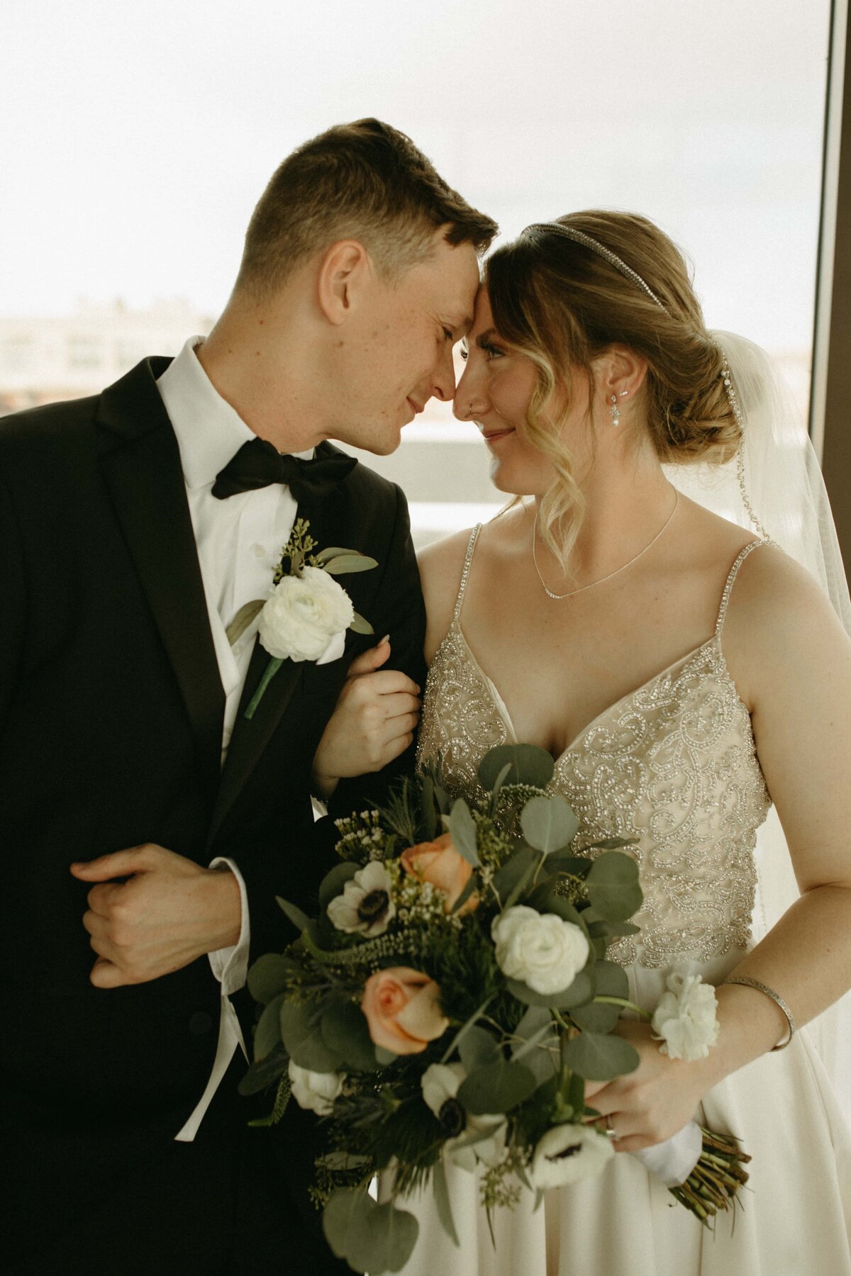 A bride and groom in wedding attire smiling at each other at an Iowa wedding, the bride holding a bouquet of white and peach flowers.