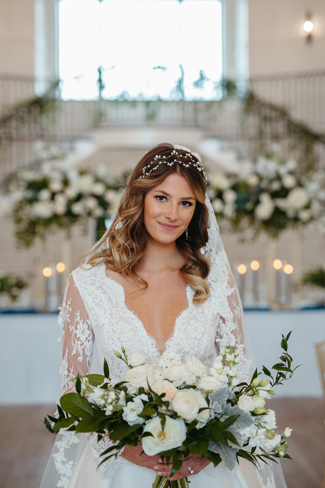A bride in a white wedding gown holing a white bouquet of flowers in front of a table with lit candles and white florals.