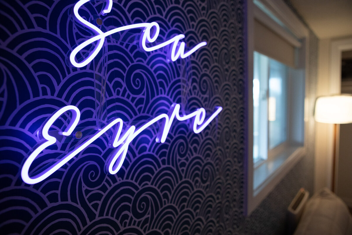 Beach house interior design details featuring a close up of a neon sign glowing with cursive writing and a patterned wallpaper with waves.
