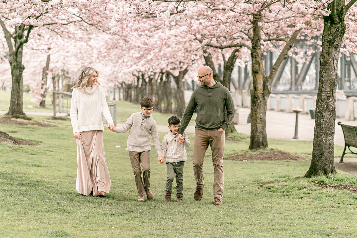 Family of four - Mom, Dad and two boys all holding hands and walking in between two rows of cherry blossom trees in Portland Oregon while the trees are in full bloom. Family is all smiling and looking at one another. Spring family portraits.