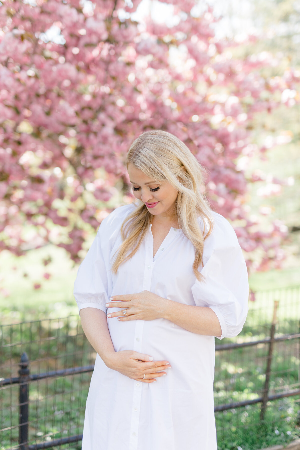 woman in white holding pregnant belly in front of cherry blossoms