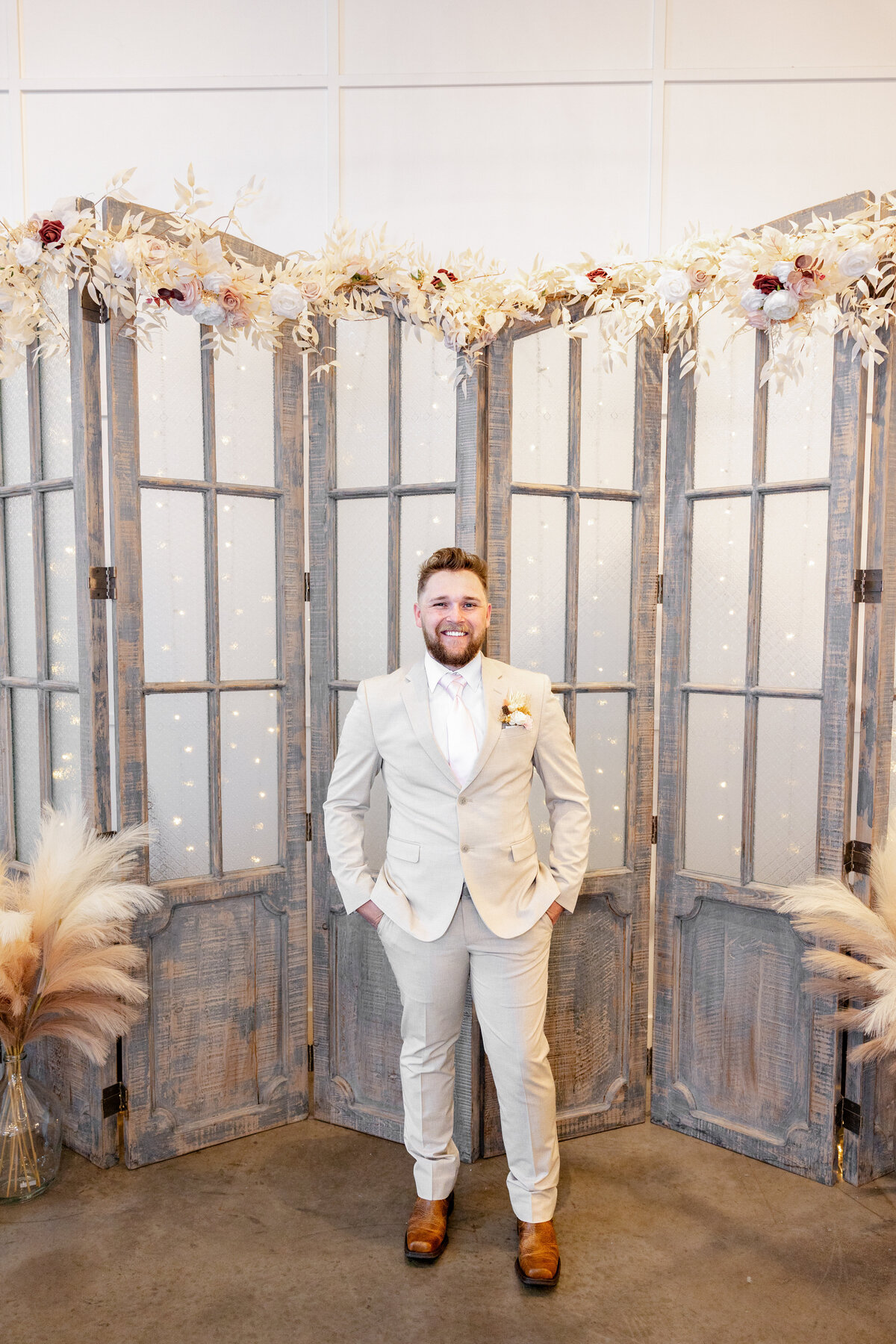 The groom, dressed in a stylish beige suit with a soft pink tie and a floral boutonniere, stands confidently in front of a rustic wooden backdrop adorned with floral accents. His smile radiates excitement and joy.