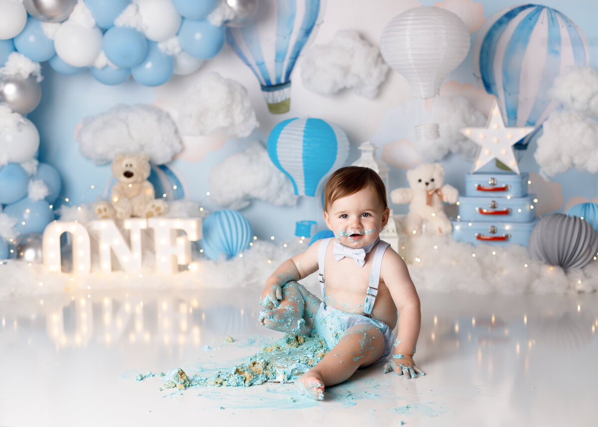 First birthday cake smash in West Palm Beach Delray Beach photography studio. Baby boy against blue and white hot air balloon and teddy bear backdrop. Baby boy is wearing suspenders and a bowtie and covered in smashed cake on his legs and arms.