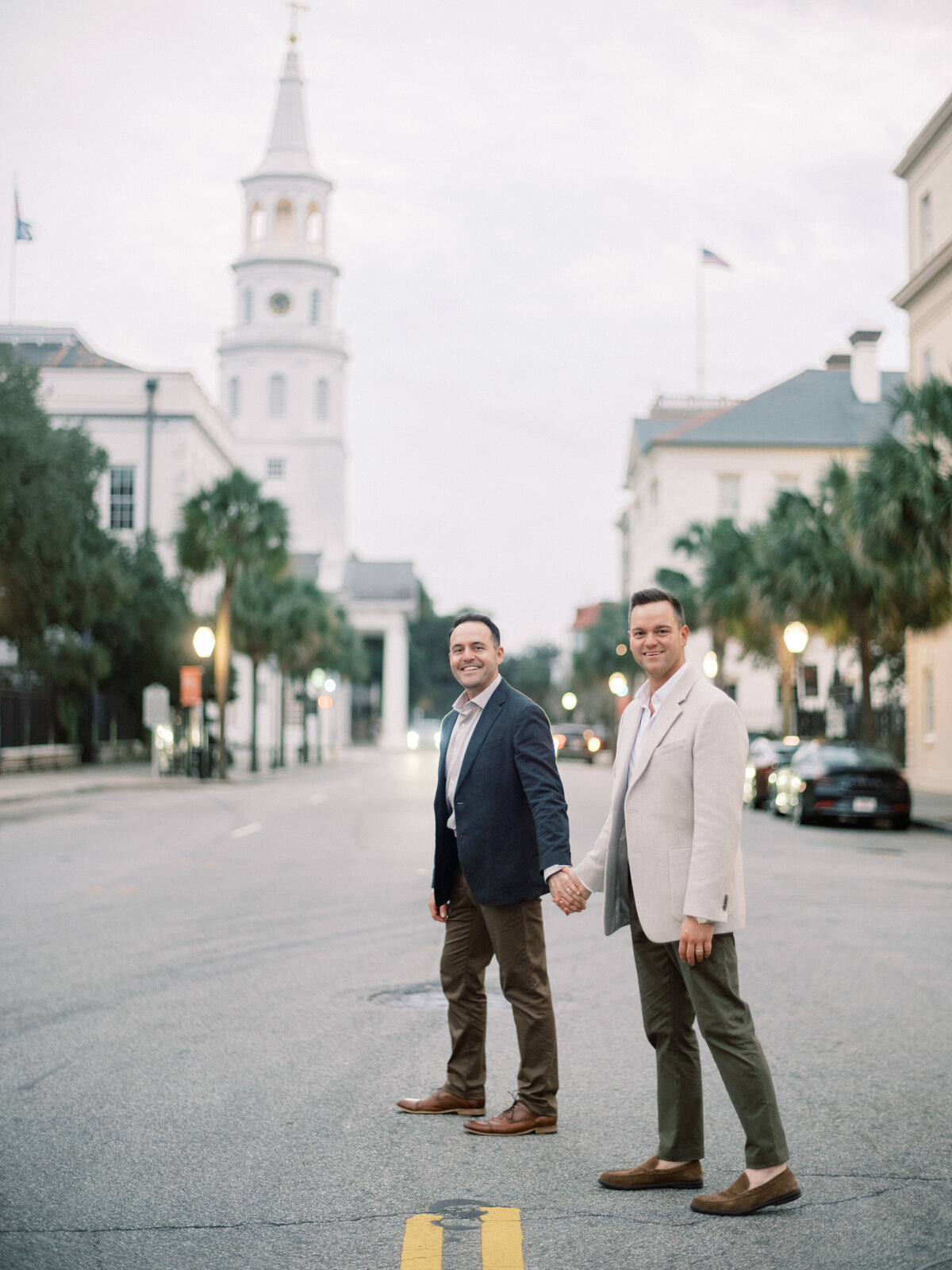 A romantic couple holds hands while walking across a charming street in Charleston, showcasing their connection against a picturesque backdrop with architecture that suggests timeless love.