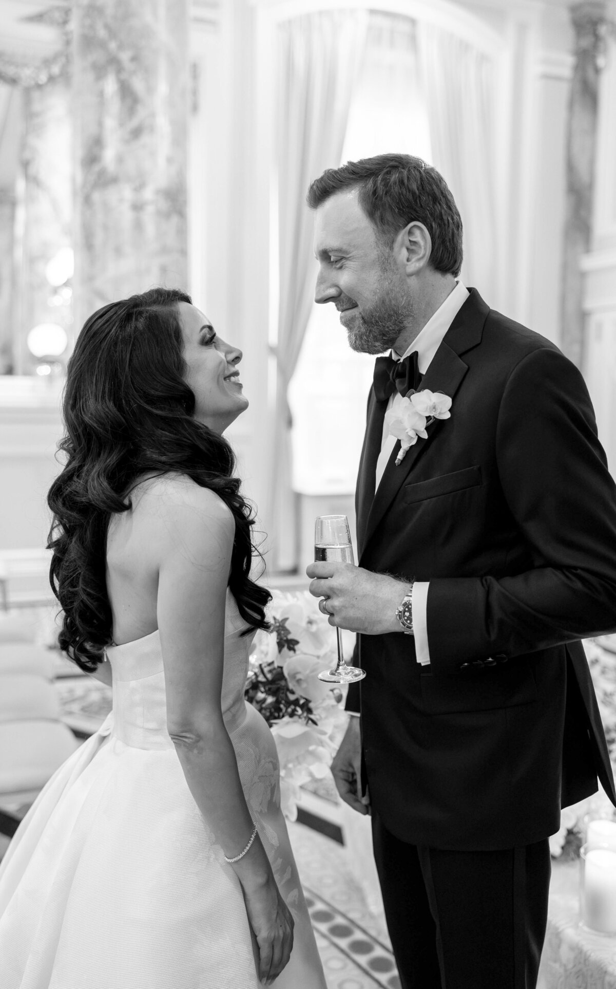 A bride and groom stand facing each other in a luxurious room. The bride wears a strapless gown and smiles at the groom, who wears a suit with a bow tie and boutonniere, holding a champagne glass. The room features ornate columns and curtains.