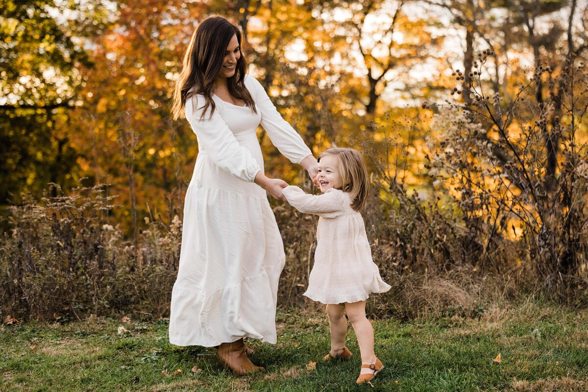 A woman in a white dress holding hands with a smiling little girl in a cream dress, both enjoying time outdoors during autumn, captured by a Pittsburgh maternity photographer.