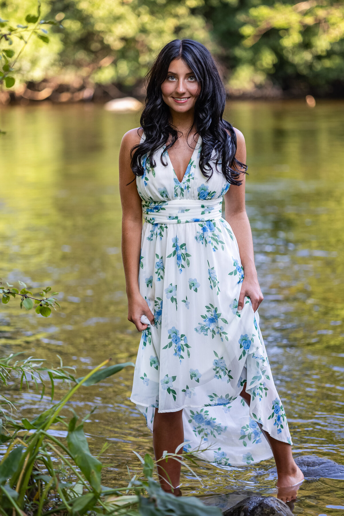 A female high school senior is posing in a long white dress with blue flowers.  She is standing in a river with water coming up to her ankles