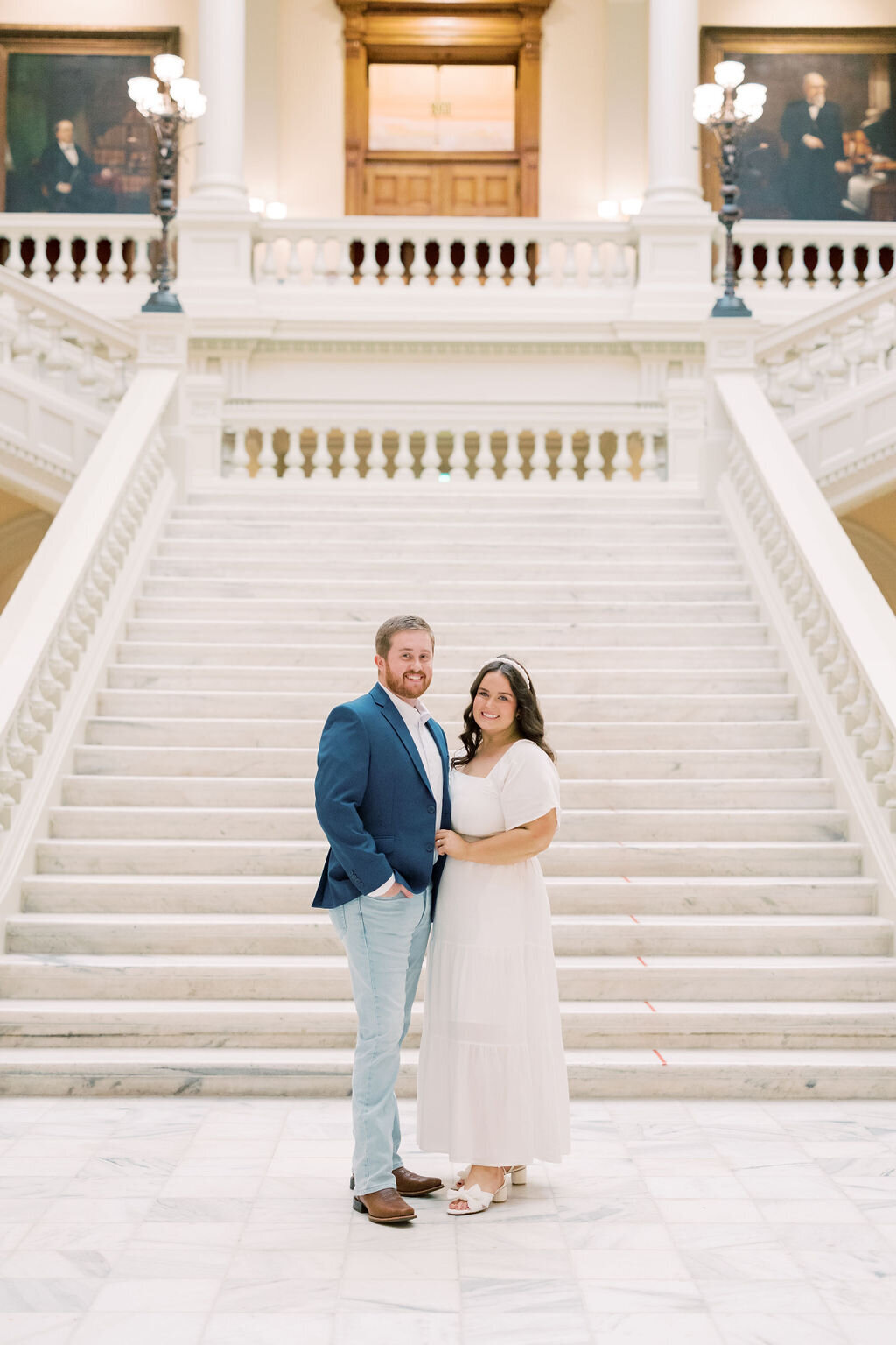 Woman in white dress holds hands with man in blue suit with Athens City Hall steps behind them