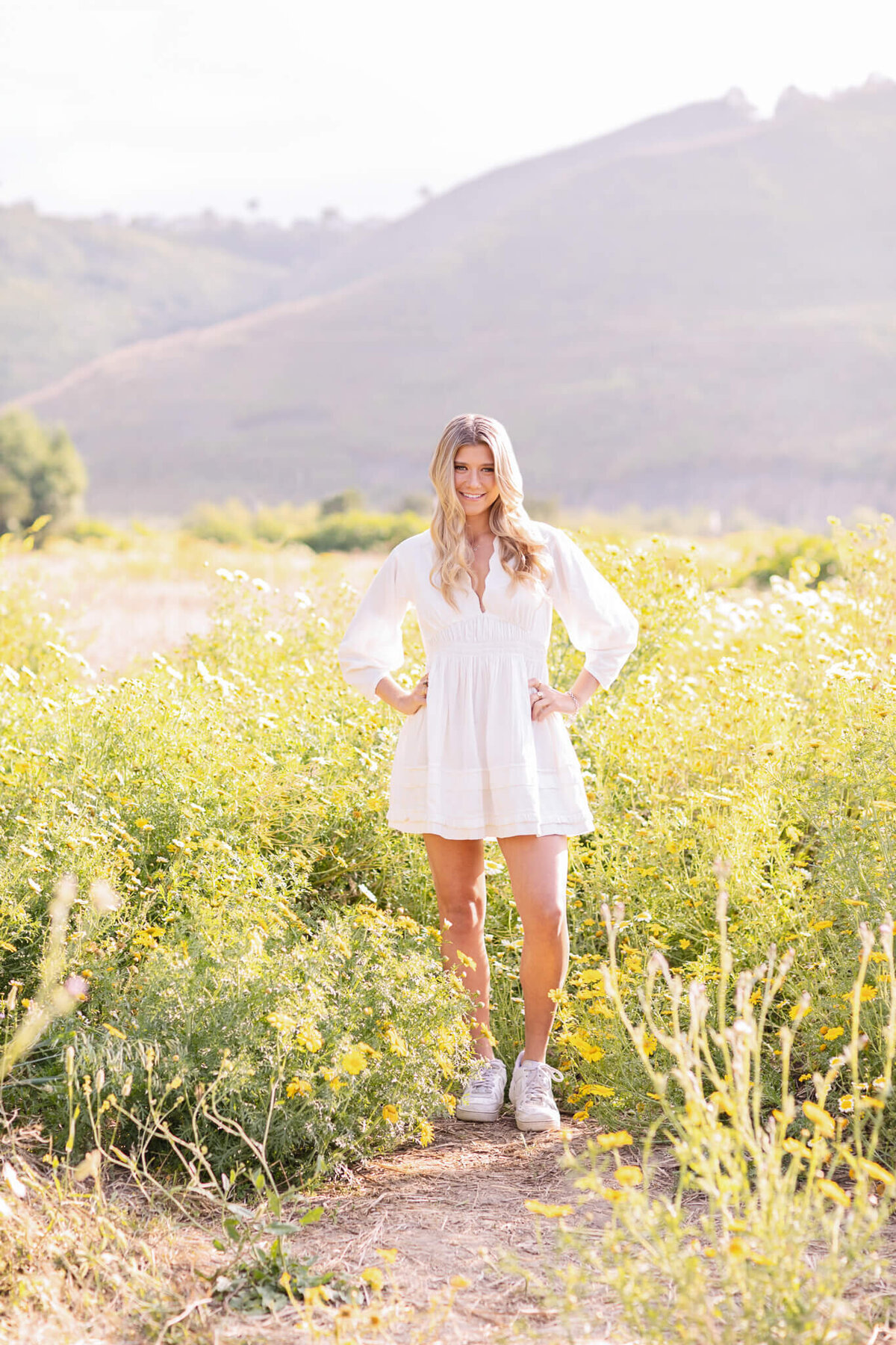 A woman in a white dress stands in a lush field with her hands on her hips. She is wearing white sneakers and has long, wavy hair. The background features rolling hills under a bright sky.