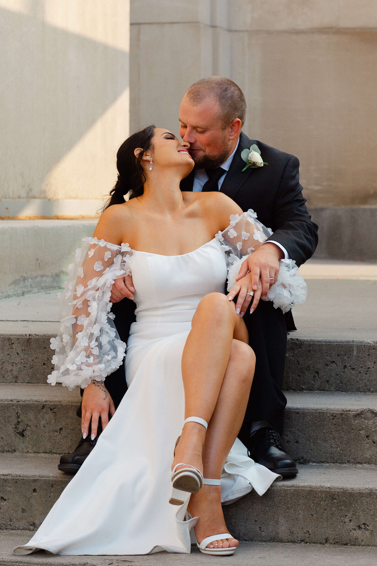 a bride and groom sitting on the stairs outside the Chateau Laurier kissing and smiling taken by Ottawa wedding photographer JEMMAN Photography