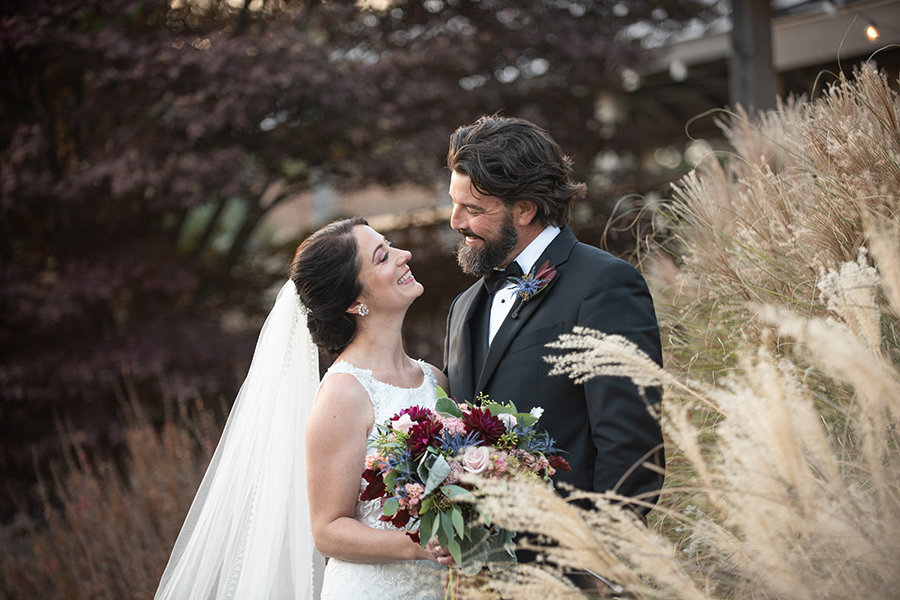 A bride and groom standing by tall grass look at each other