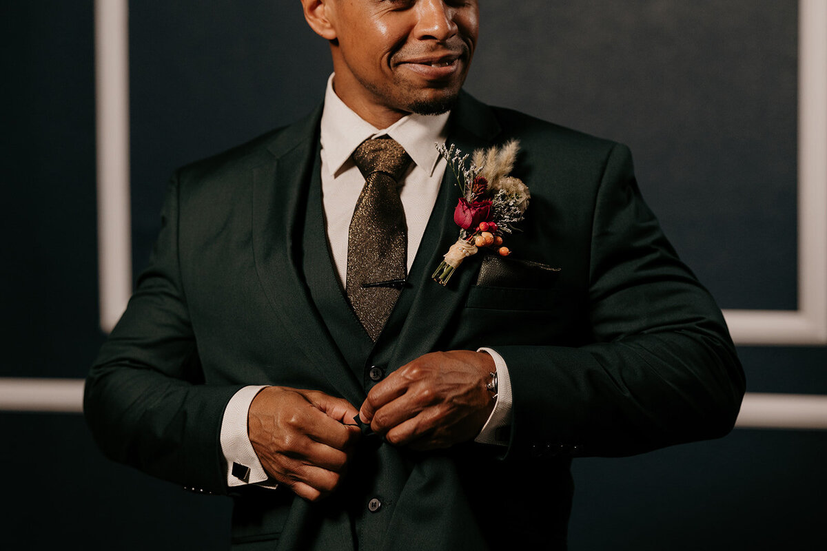 The groom, dressed in a dark green suit with a rustic boutonniere of dried flowers and berries, adjusts his jacket with a confident expression.