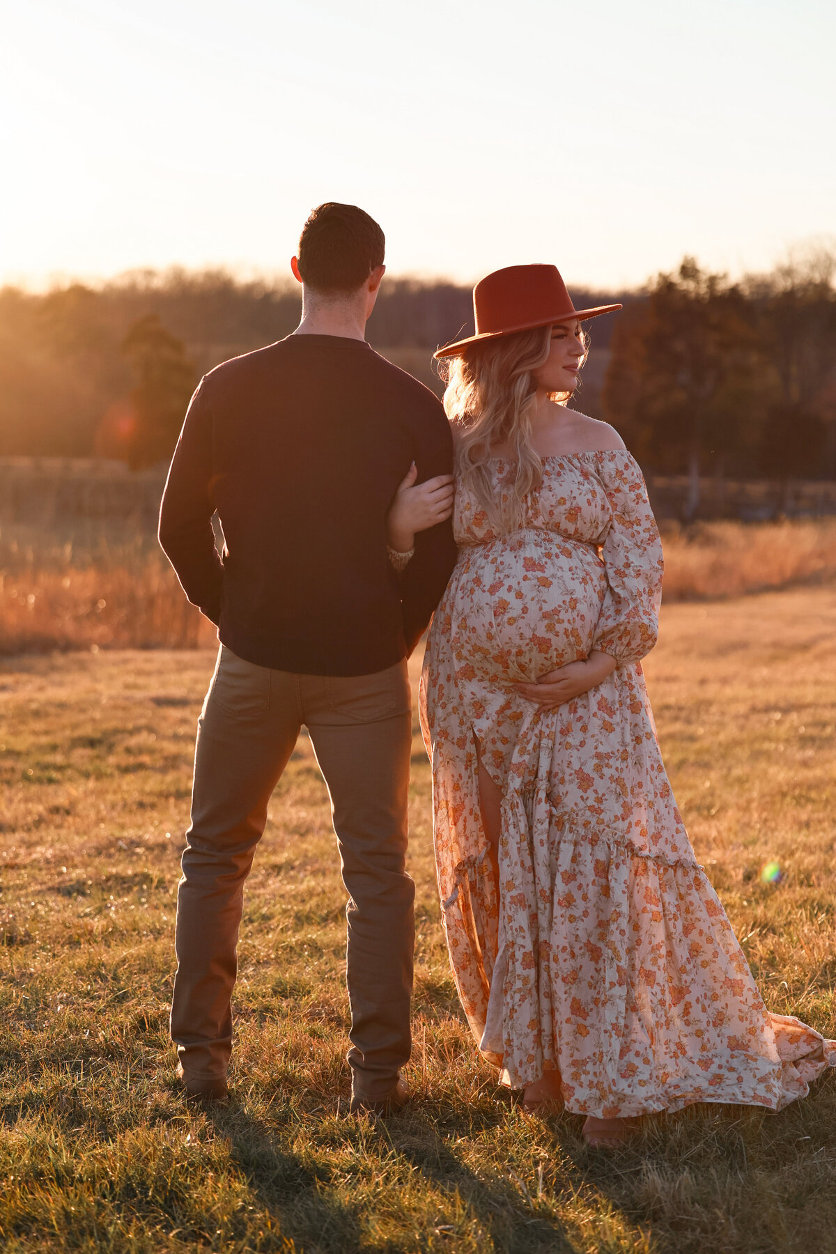 pregnant woman holding her husbands arm while holding her pregnant belly while standing in a field at sunset