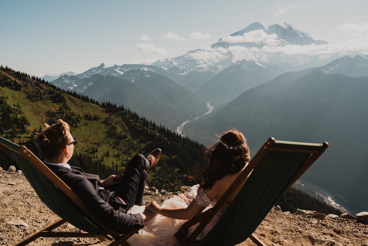 after learning how to elope in Washington State, a couple enjoys the sunset behind Mount rainier as they  hold hands sitting in lounge chairs