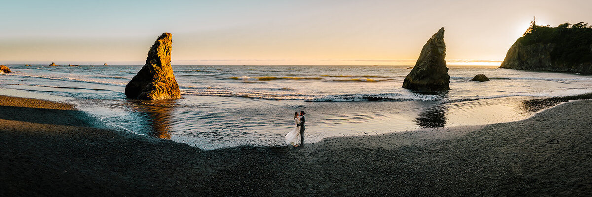 A sweeping panoramic of a rocky beach with a couple in their wedding attire perfectly centered between two triangular sea stacks. Taken from a higher perspective and through the eyes of their adventure elopement photographers, we see the couple embrace at the waterline, where the gentle ocean waves meet the rocky, pebble strewn beach. Sunset colors paint the sky and light the sea stacks with a golden hugh.