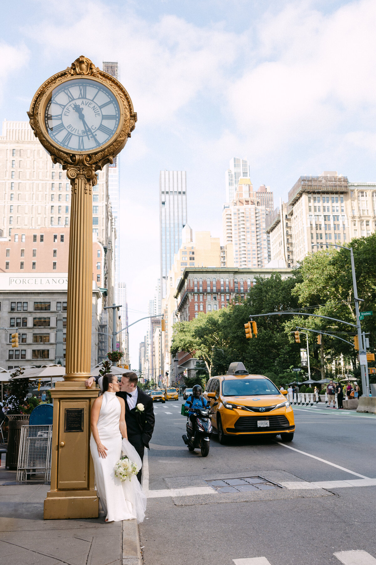 Bride and groom sharing a kiss under the iconic gold Fifth Avenue clock near Central Park, with NYC traffic and yellow taxis passing by in the background

