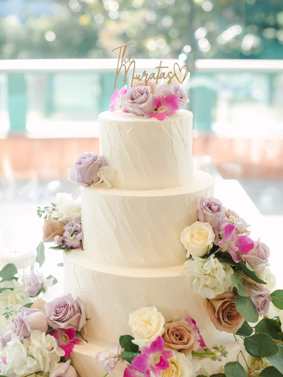 A three-tiered white wedding cake is adorned with pastel roses, orchids, and eucalyptus leaves. The top has a heart-shaped topper with text.